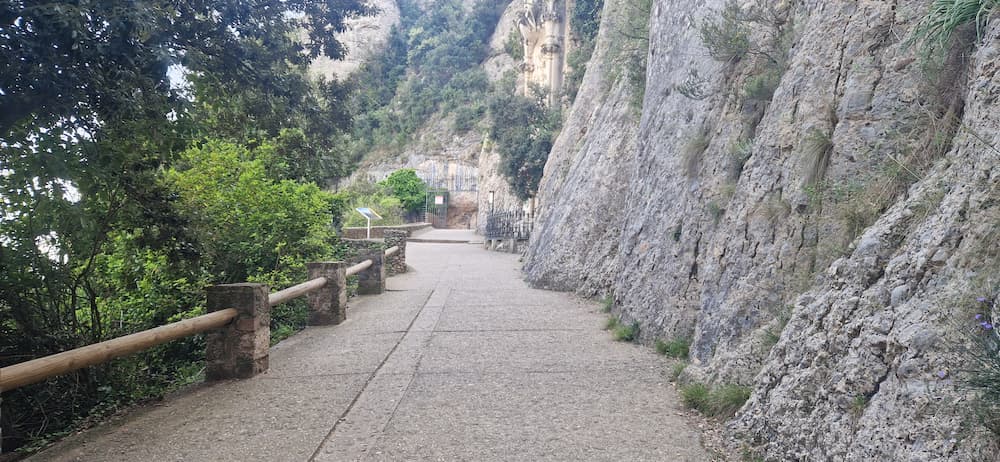 Santa Cova path arrival area in Montserrat, with paved walkway, rock wall and railings near the chapel entrance